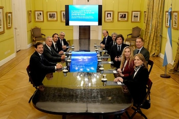 Grupo de personas sentadas alrededor de una mesa de reuniones oscura. Hay una pantalla mostrando texto, una bandera de Argentina y cuadros en las paredes