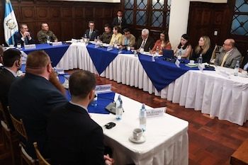 Sala de reuniones formal con paneles de madera; varias personas sentadas en mesas con manteles blancos y azules. Una bandera de Guatemala y botellas de agua son visibles
