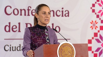 Claudia Sheinbaum habla ante dos micrófonos en un podio de madera con el escudo nacional de México sobre un fondo con letras rojas.