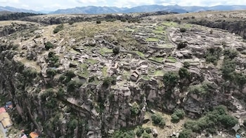 Vista aérea de una antigua ciudadela de piedra en una colina rocosa con estructuras circulares y rectangulares, rodeada de vegetación y montañas