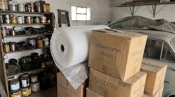 Un rollo de plástico de burbujas y cajas de cartón escritas ante estanterías con latas y un automóvil cubierto con una lona.