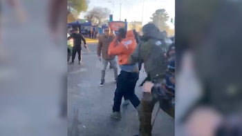 Um grupo de pessoas na rua, uma delas com um vestido laranja. Outra pessoa vestindo um casaco escuro e máscara. Muitas árvores e estruturas