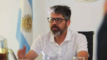 Hombre barbudo con anteojos y camisa blanca estampada, gesticulando en una oficina con la bandera argentina visible detrás
