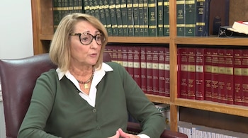 Una mujer con gafas y suéter verde habla sentada frente a una biblioteca repleta de libros jurídicos de encuadernación roja y verde
