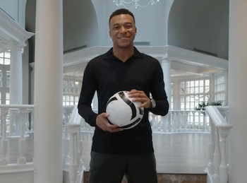 Kylian Mbappé, un hombre joven sonriente, de pie en un vestíbulo blanco con columnas y pasamanos ornamentados, sostiene un balón de fútbol