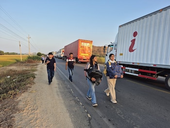 Vista lateral de personas caminando por el arcén de una carretera. A su derecha, una larga fila de camiones y autobuses está detenida. Al fondo, campos y colinas
