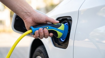 Mano inserta un enchufe de colores azul, verde y negro con un cable amarillo en el puerto de un automóvil blanco. El enchufe muestra indicadores de carga iluminados.