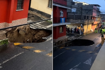 Un gran socavón en el asfalto de una calle junto a una edificación roja con una bandera de Guatemala y personas en la acera.