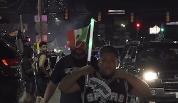 Vista nocturna de varias personas, algunas con camisetas de los San Antonio Spurs, reunidas cerca de vehículos en una calle iluminada con luces de tráfico y de vehículos