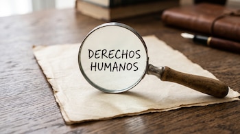 Una lupa sobre un papel con el texto Derechos Humanos, junto a una pluma y un cuaderno sobre una mesa de madera.
