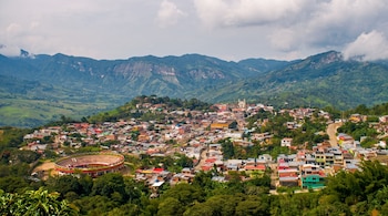 Vista elevada de un pueblo con casas, una plaza circular, árboles y montañas al fondo bajo un cielo nublado.