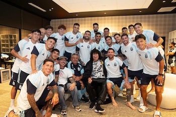 Un grupo grande de hombres posa para una foto. Muchos visten camisetas de fútbol azul claro y blanco. Mona Jiménez está sentado en el centro junto a Lionel Messi