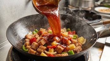 Un cucharón vierte salsa agridulce rojiza sobre trozos de cerdo, piña, pimientos y cebolla en un wok humeante. Se observa vapor ascendiendo del wok.