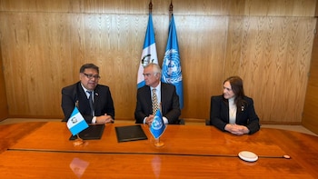 Tres funcionarios, dos hombres y una mujer, sentados en una mesa de madera con banderas de Guatemala y la ONU al fondo, y pequeñas banderas de escritorio