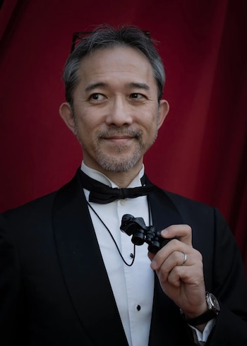 Un hombre de cabello gris y barba viste esmoquin, camisa blanca y corbata de lazo. Sostiene unos prismáticos de ópera y lleva un anillo y un reloj