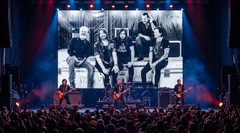 Músicos interpretan un concierto en un escenario iluminado frente al público, con una pantalla al fondo que muestra una foto en blanco y negro.