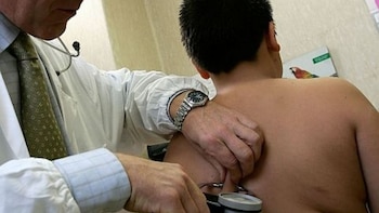 Un médico en bata blanca con estetoscopio examina la espalda de un niño sin camiseta. Parte de una imagen con un loro es visible al fondo