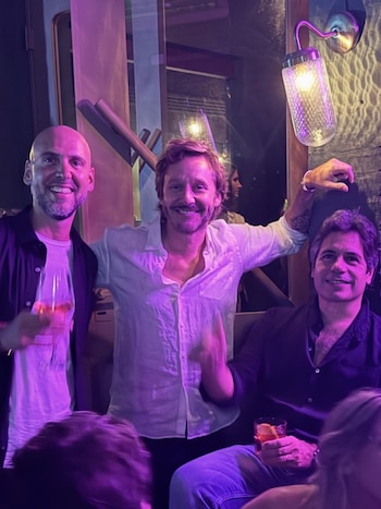 Tres hombres sonríen en una fiesta con luces moradas; uno calvo sostiene una copa, el del medio viste camisa blanca y el tercero, sentado, sostiene un cóctel