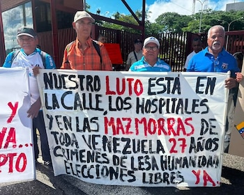 Varios hombres sostienen una gran pancarta blanca con texto manuscrito en letras negras y rojas frente a una reja metálica