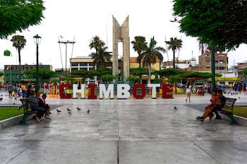 Vista de la Plaza Mayor de Chimbote con un gran letrero de 'CHIMBOTE' en letras rojas y blancas, un monumento central, palmeras, y personas sentadas en bancos