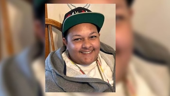 Persona joven que sonríe con gorra de béisbol negra, chaqueta gris con capucha y camiseta blanca, sentada en una silla de madera
