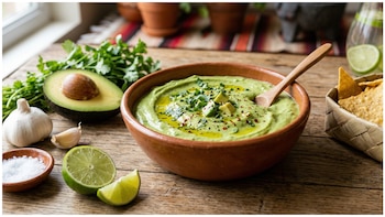Un tazón de dip de aguacate verde cremoso con aceite, cilantro y chiles, junto a aguacate, ajo, lima, sal y totopos en canasta sobre madera