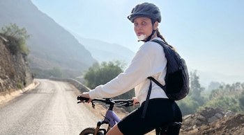 Anna Carina Copello montando una bicicleta de montaña en una carretera rodeada de montañas, lleva casco, mochila y un protector facial.