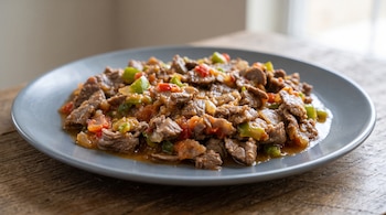 Un plato gris redondo con tiras de carne cocida en salsa con pedazos de tomate y pimiento sobre una mesa de madera.