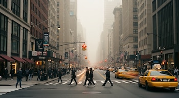 Vista de una avenida concurrida en una ciudad de Estados Unidos. Varias personas cruzan la calle, taxis amarillos circulan y altos edificios se alzan bajo un cielo brumoso.
