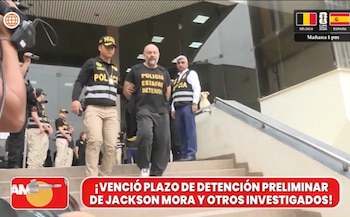 Hombre con camiseta negra que dice "POLICIA ESTAFAS DETENIDO" es escoltado por dos agentes policiales que llevan chalecos reflectantes. Otros agentes, una cámara de televisión y un edificio son visibles