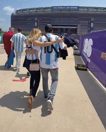 Hombre y mujer caminando abrazados. Él con camiseta de fútbol argentina número 10. Ella con blusa de rayas. Estadio con cartel FIFA World Cup 2026