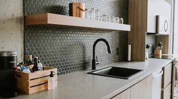 Fregadero negro con grifo curvo en una encimera gris, bajo un estante de madera con vasos y un muro de azulejos hexagonales oscuros.
