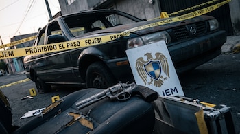 Automóvil negro, cinta amarilla FGJEM, letrero con emblema FGJEM, pistola en asiento, marcadores de evidencia.