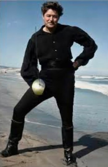 Un hombre con camisa, pantalón y botas negras sostiene una espada sobre la arena frente al mar