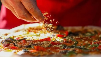 Primer plano de una mano esparciendo pequeñas piezas claras sobre una pizza con ingredientes rojos y verdes, sobre un fondo rojo difuminado