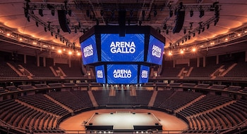 Vista aérea del interior vacío de Arena Gallo, con filas de asientos oscuros, un escenario central iluminado y pantallas LED octogonales mostrando el logo 'Arena Gallo'.