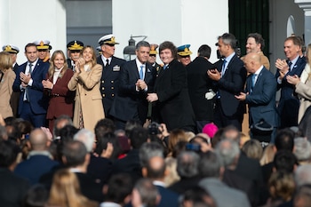 Un grupo de hombres en traje y uniforme militar, junto a mujeres en abrigos, aplauden de pie frente a una fachada blanca, con una multitud desenfocada en primer plano