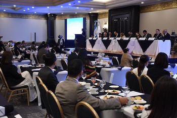 Vista panorámica de un salón con mesas redondas ocupadas por asistentes, un podio, una pantalla grande, una mesa principal con oradores y banderas