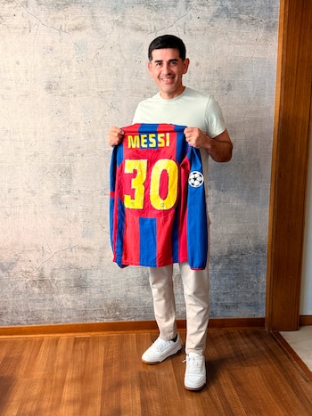 Hombre sonriente, camiseta de fútbol azul y roja con el nombre Messi y el número 30, parche de balón de fútbol, pantalón claro, zapatillas blancas, pared gris, suelo de madera