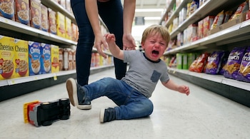 Niño rubio con camiseta de rayas llora sentado en el suelo de un supermercado. Un adulto se inclina cerca y un camión de juguete rojo y amarillo está volcado.