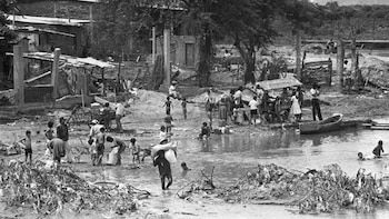 Fotografía en blanco y negro muestra personas, incluyendo niños, transitando por una zona inundada con barro y escombros. Se ven casas dañadas y un bote
