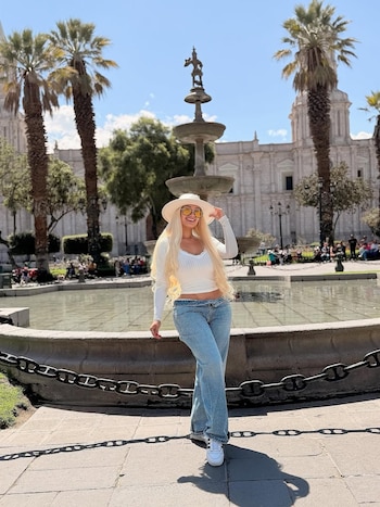 Mujer rubia con sombrero blanco y gafas de sol amarillas posa frente a una fuente ornamental en una plaza pública con palmeras y edificios al fondo