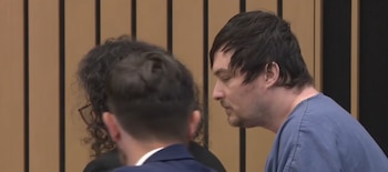 Un hombre caucásico de pelo oscuro y barba incipiente, con una camiseta azul de manga corta, mira hacia abajo en una sala
