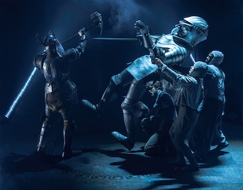 Escena teatral oscura con dos figuras con armadura; una grande y plateada es atacada por otra oscura con martillo y bastón luminoso. Tres figuras rodean al caballero grande