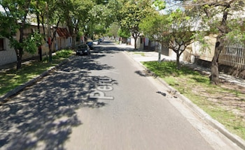 Calle asfaltada "Perú" con árboles a los lados, sombras de copas sobre la vía, casas de baja altura, autos estacionados y aceras de cemento