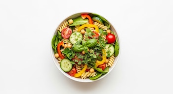 Bowl blanco con pasta fusilli, espinacas, rodajas de pepino, pimientos rojos y amarillos, tomates cherry, garbanzos y hierbas frescas.