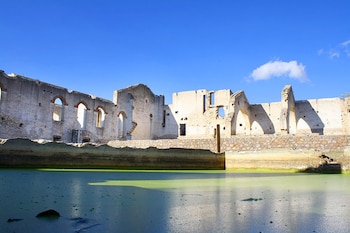 Ruinas de una antigua construcción de piedra y argamasa con arcos y paredes parciales, reflejadas en un cuerpo de agua verde bajo un cielo azul claro