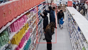 Toma de cámara de seguridad de un pasillo de supermercado con estantes de champú y varias personas caminando entre los exhibidores