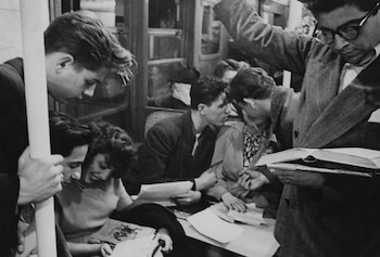 Fotografía en blanco y negro de varias personas dentro de un vagón de metro; algunas de pie y otras sentadas, leyendo periódicos y libros, o conversando