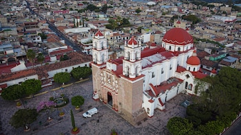 Vista aérea de una gran iglesia blanca con tejado y cúpulas rojas, situada en una plaza adoquinada con un vehículo blanco y árboles frondosos, rodeada por un denso pueblo